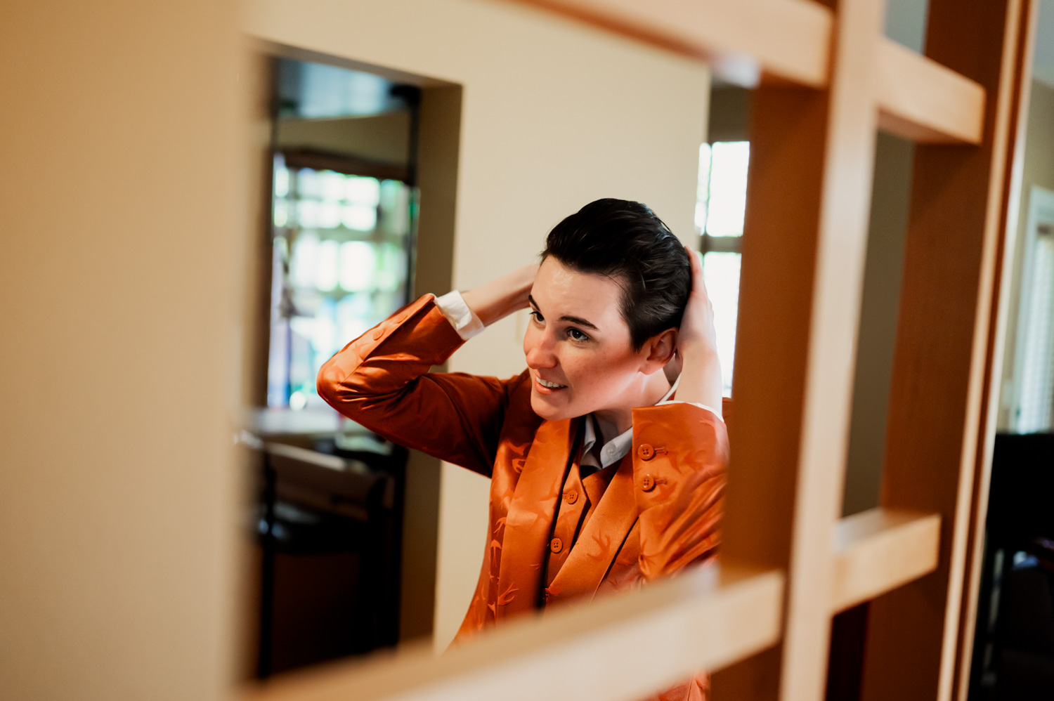 A person in an orange jacket stands in front of a mirror, smiling while adjusting their hair. A wedding at the Hoyt Arboretum Redwood Deck is mentioned as their destination.