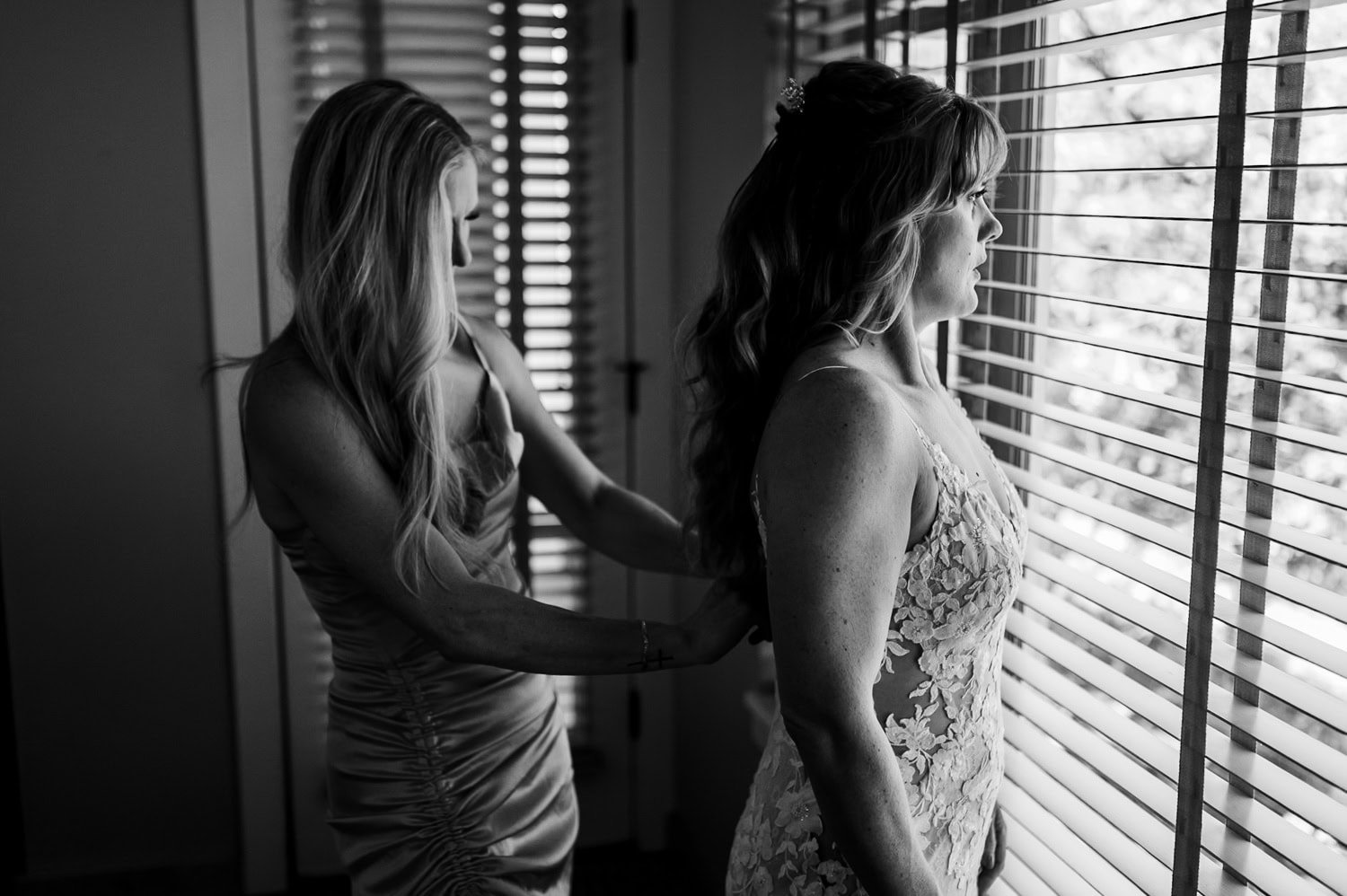 A bride in a lace dress stands by a window as another woman assists with the zipper, preparing for the wedding at Hoyt Arboretum Redwood Deck.