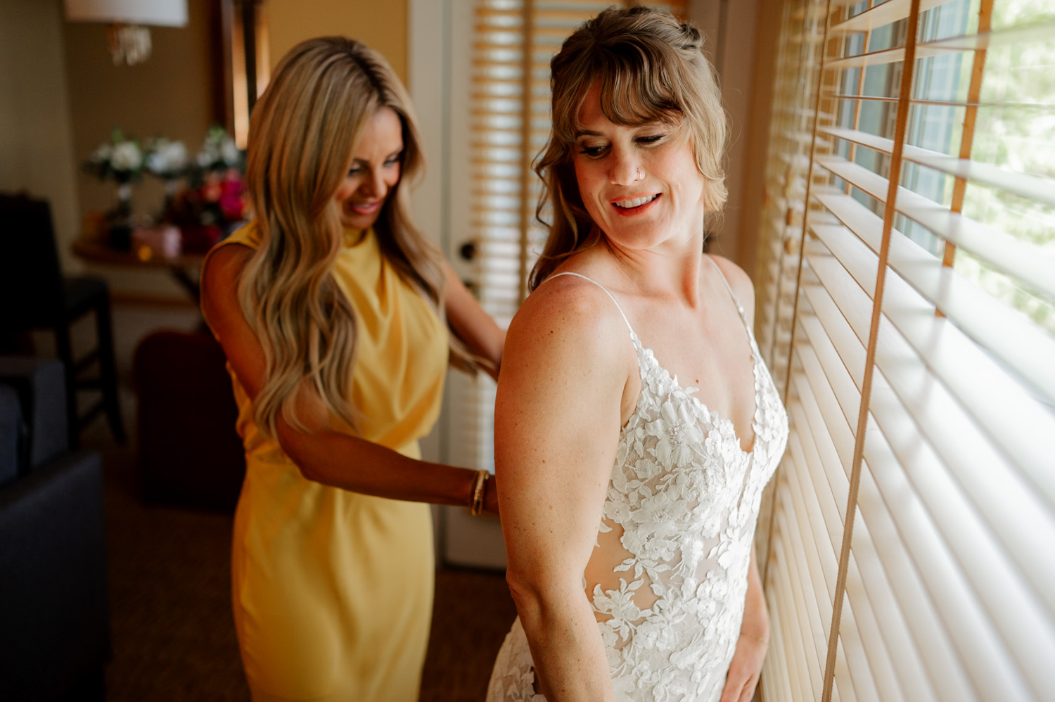 Two women stand together in a room as they prepare for a wedding at the Hoyt Arboretum Redwood Deck. The bride, wearing a bridal gown, smiles brightly, while the woman next to her in a yellow dress adjusts her outfit near the window blinds.