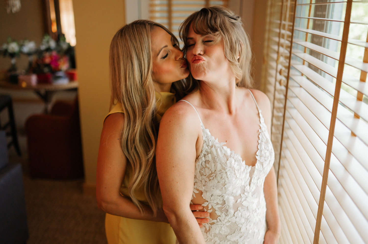 Two women, one wearing a yellow dress and the other in a white dress, smiling and posing playfully near window blinds.