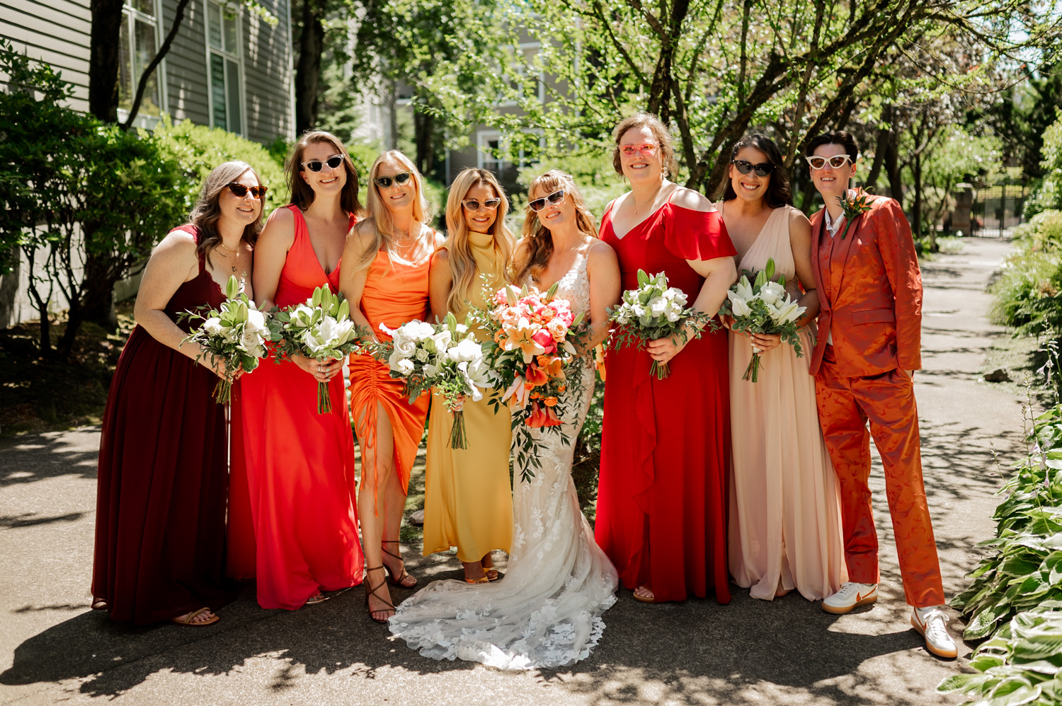 A bride in a white gown poses at the Hoyt Arboretum Redwood Deck, surrounded by seven bridesmaids in colorful dresses and suits. Each bridesmaid holds a bouquet as they join in celebration before the wedding ceremony. The lush greenery of the arboretum surrounds them, creating a serene and joyful atmosphere.
