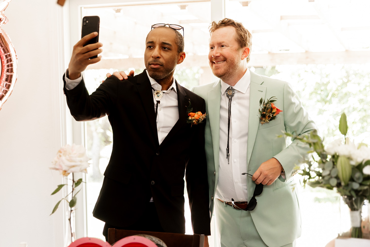Two men in suits smile brightly as they take a selfie indoors with abundant natural light. They are preparing for a wedding at the Hoyt Arboretum Redwood Deck.