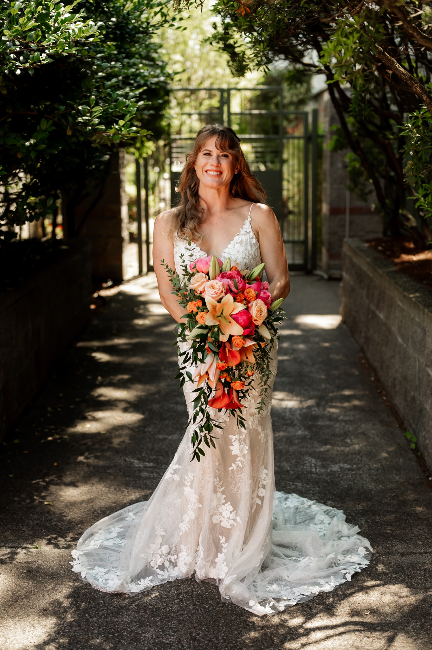 Bride in a white lace gown holds an orange and pink flower bouquet on a shaded garden path at Hoyt Arboretum Redwood Deck.