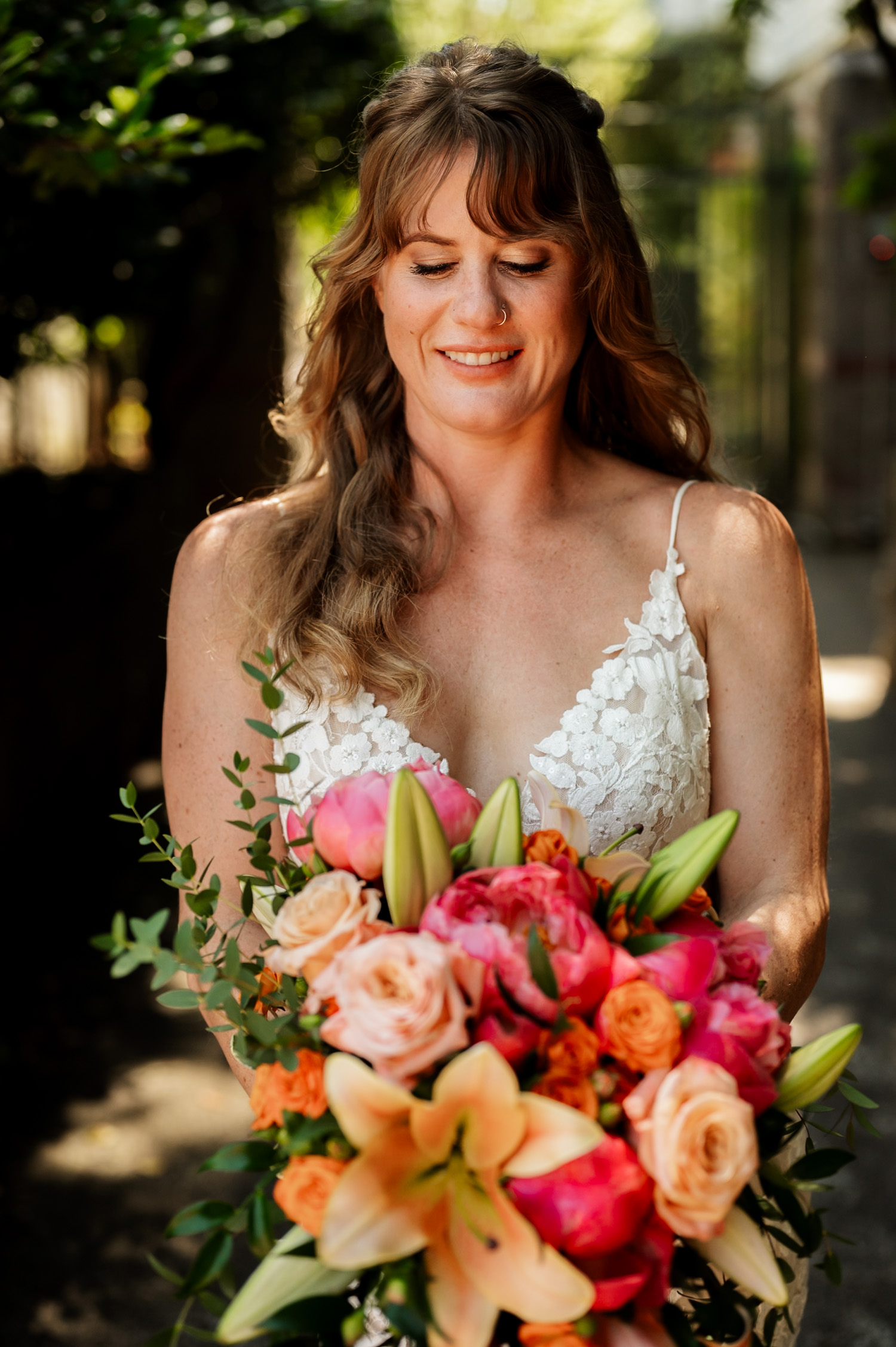 The bride smiles while holding a vibrant bouquet of pink lilies, roses, and lush greenery against the backdrop of trees at the Hoyt Arboretum Redwood Deck.