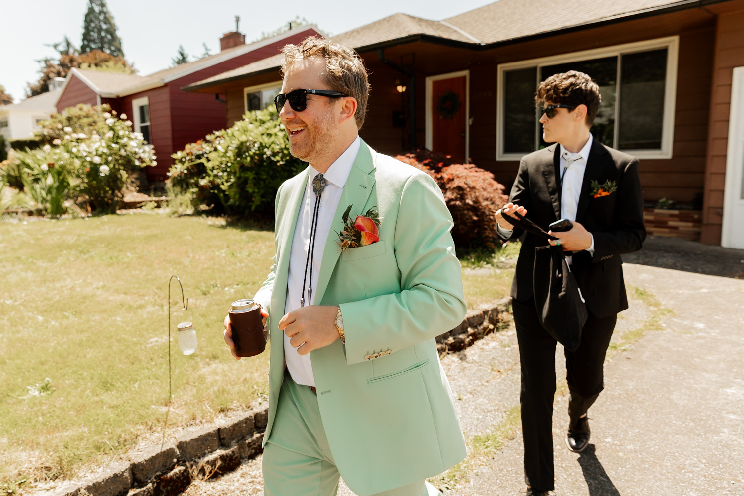 Two men in suits, one in mint green holding a drink and the other in black, walk together outside on a sunny day at the Hoyt Arboretum Redwood Deck, preparing for a wedding.