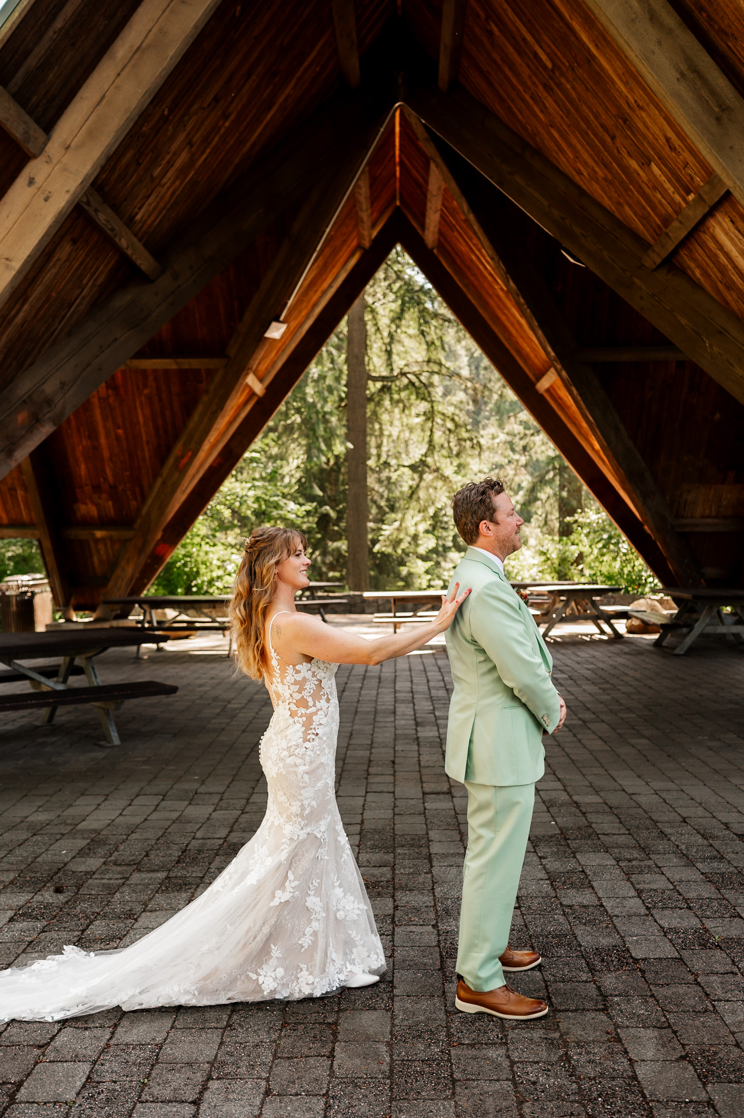 In the rustic wooden pavilion at Hoyt Arboretum, the bride in a lace gown gently touches the grooms shoulder during their first look, capturing a tender moment for wedding photos.
