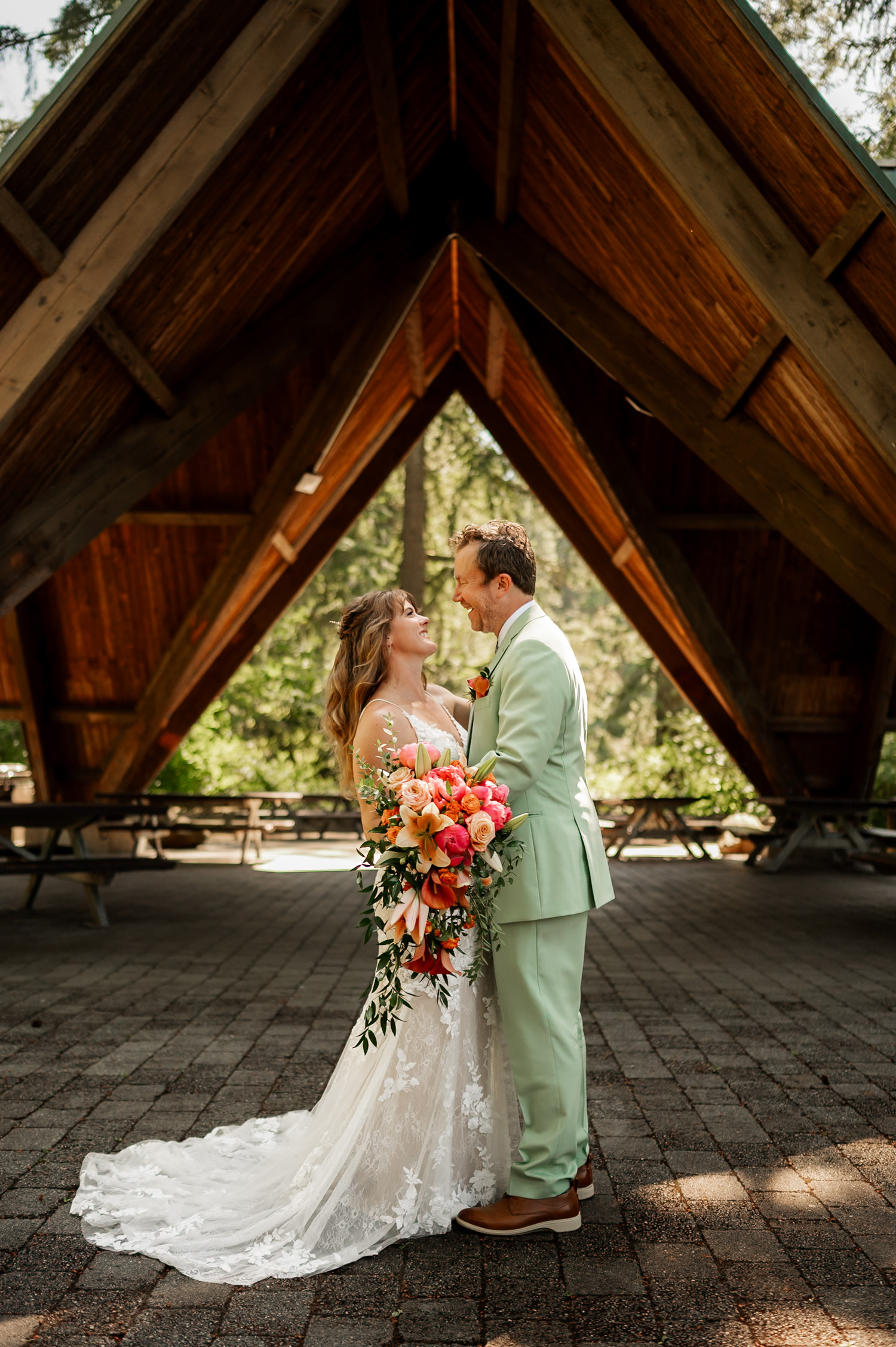 Bride and groom smiling broadly under a wooden arch at Stevens Pavilion, Hoyt Arboretum. The bride holds a colorful bouquet of flowers, capturing a joyful moment in their wedding photos.
