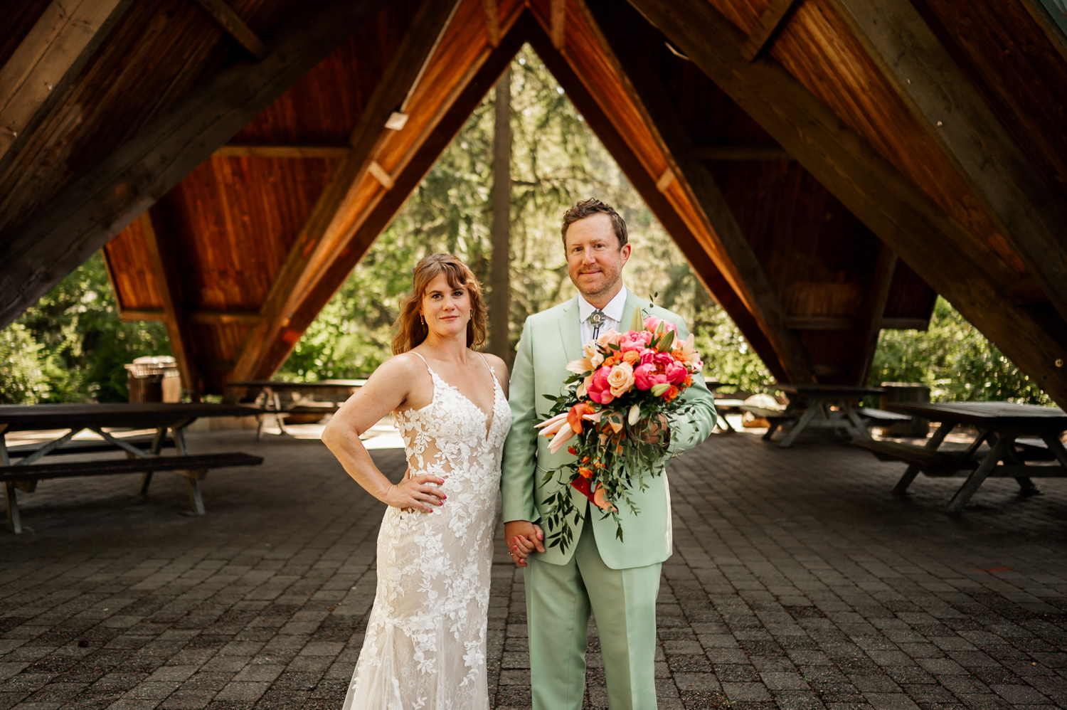 Bride in a lace dress and groom in a mint suit hold flowers while standing under the wooden structure at Hoyt Arboretums Stevens Pavilion.