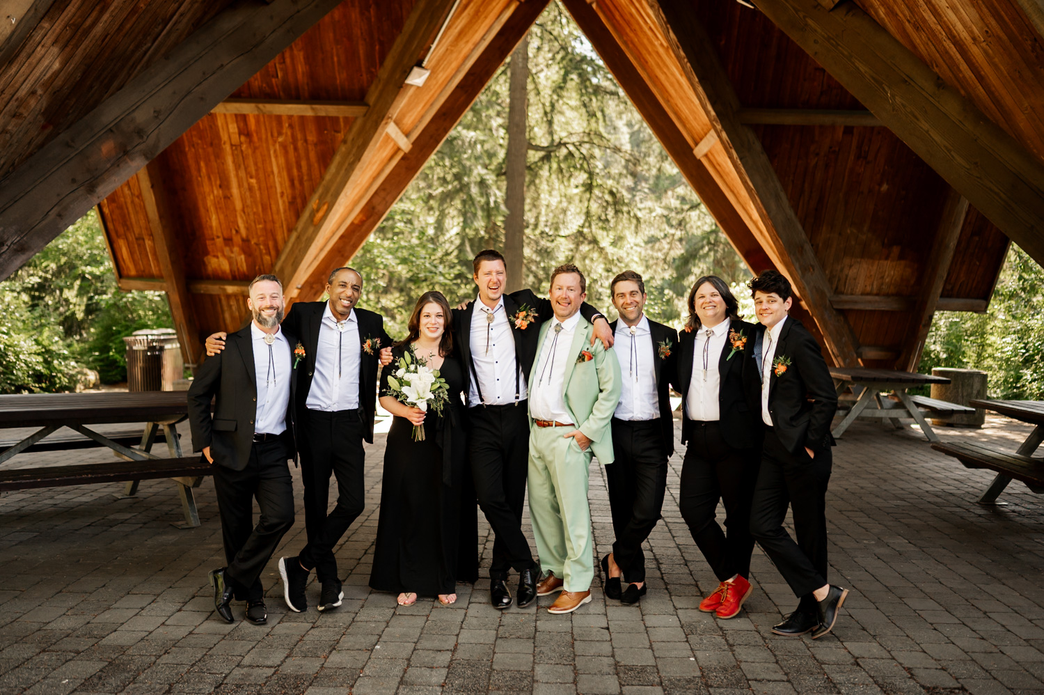 A group of people in formal attire pose together under Stevens Pavilion at Hoyt Arboretum, capturing wedding photos with lush greenery in the background.