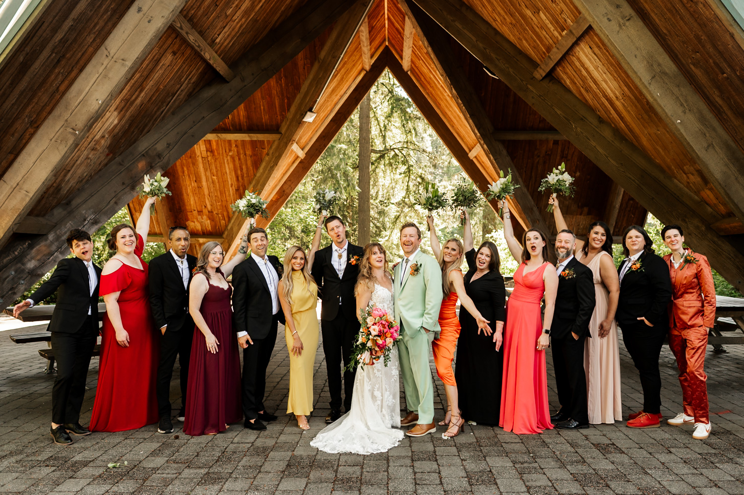 Bride and groom with a vibrant wedding party under the wooden structure at Stevens Pavilion, Hoyt Arboretum.