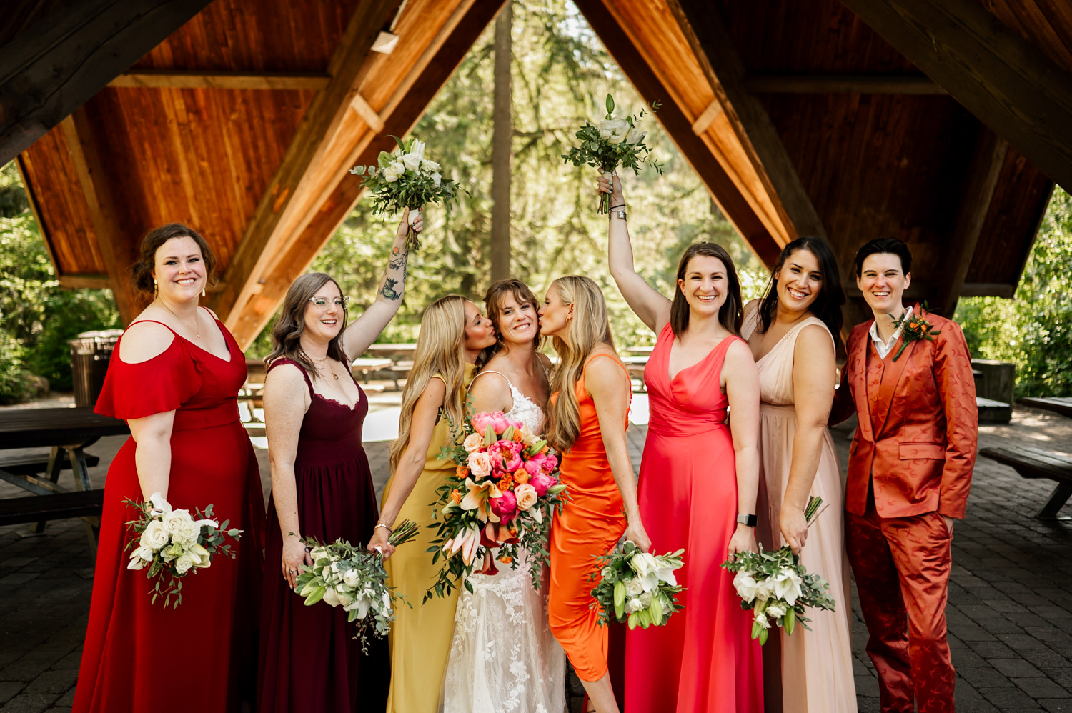 A bridal party in colorful dresses smiling and raising their bouquets under a wooden pavilion at Hoyt Arboretum Stevens Pavilion, surrounded by greenery.