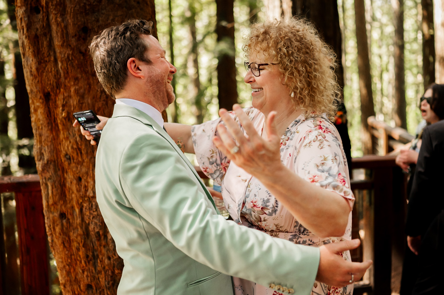 Two people smiling and hugging in a forest setting, both dressed in light-colored clothes, celebrating at the Redwood Deck in Hoyt Arboretum.