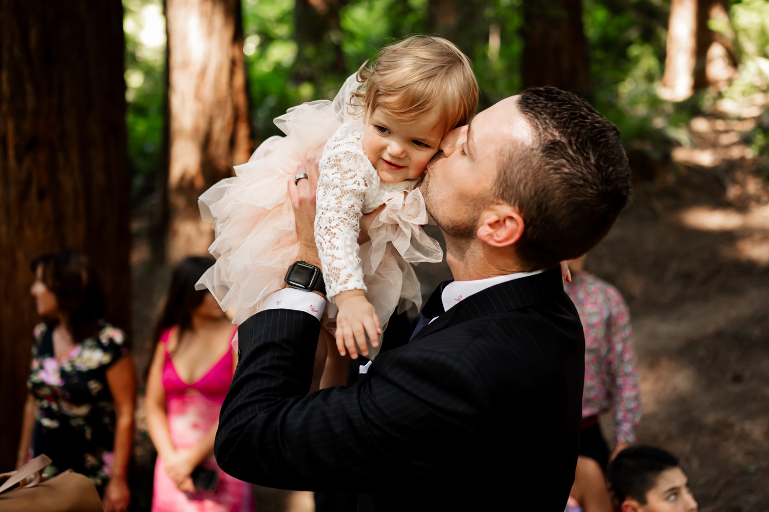 A man in a suit kisses a smiling toddler in a lace dress against the forest backdrop of the Redwood Deck at Hoyt Arboretum. Guests are mingling joyfully in the background, capturing a moment from a wedding ceremony.