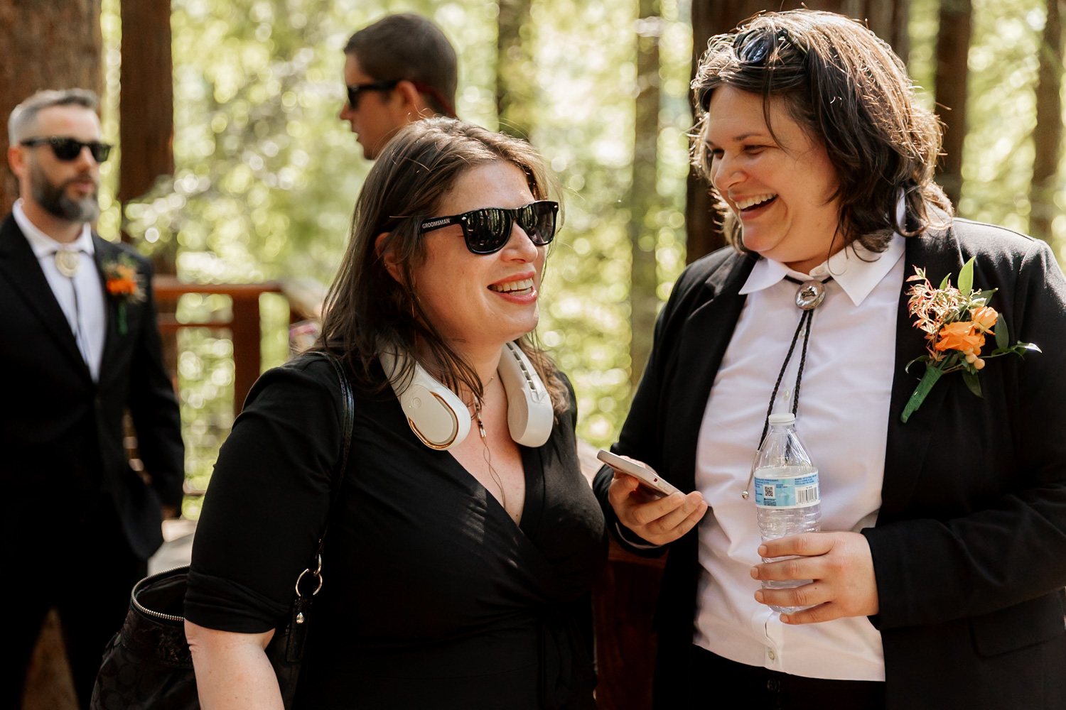Two people dressed in black clothing smile and talk to each other outdoors during a wedding ceremony. They stand among tall redwood trees at the Redwood Deck in Hoyt Arboretum, with sunlight streaming through the branches. Other guests can be seen mingling in the background.