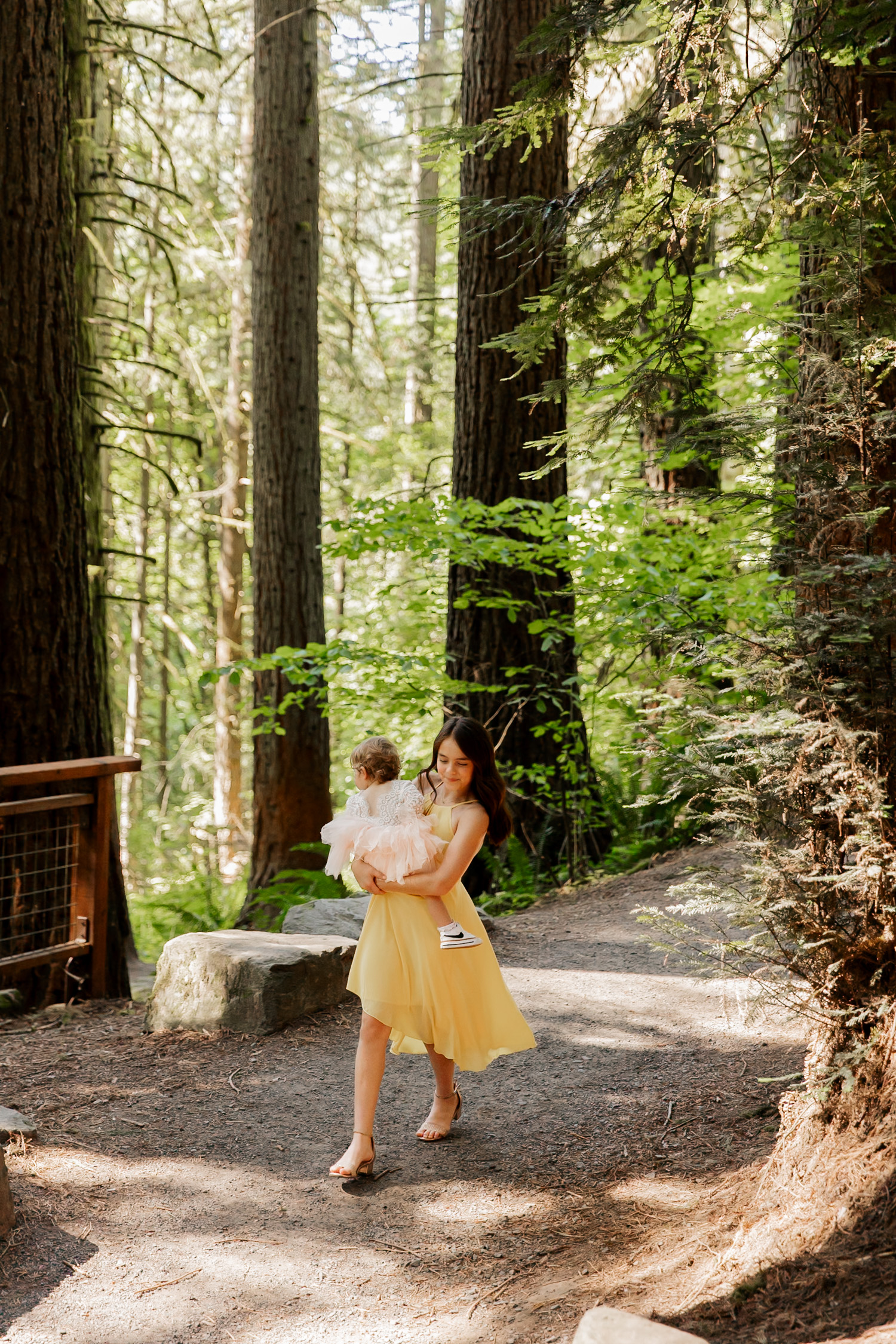 A woman in a yellow dress holds a baby as she walks along a forest path, surrounded by towering trees.