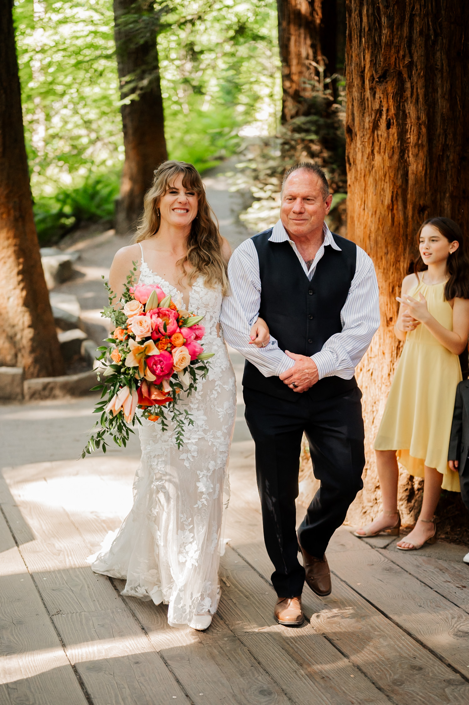 A bride holding a bouquet walks down a forest path with an older man at the Redwood Deck in Hoyt Arboretum. A young girl in a yellow dress stands nearby amidst the trees.
