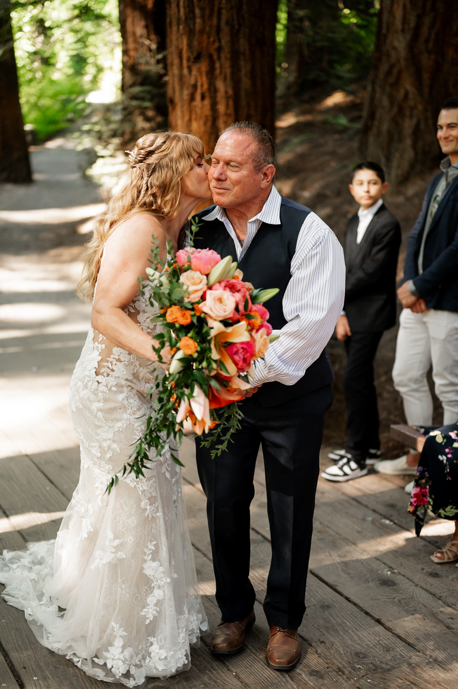 A bride kisses a smiling older man in a wooded area at the Redwood Deck in Hoyt Arboretum, holding a vibrant bouquet, with several onlookers standing nearby.