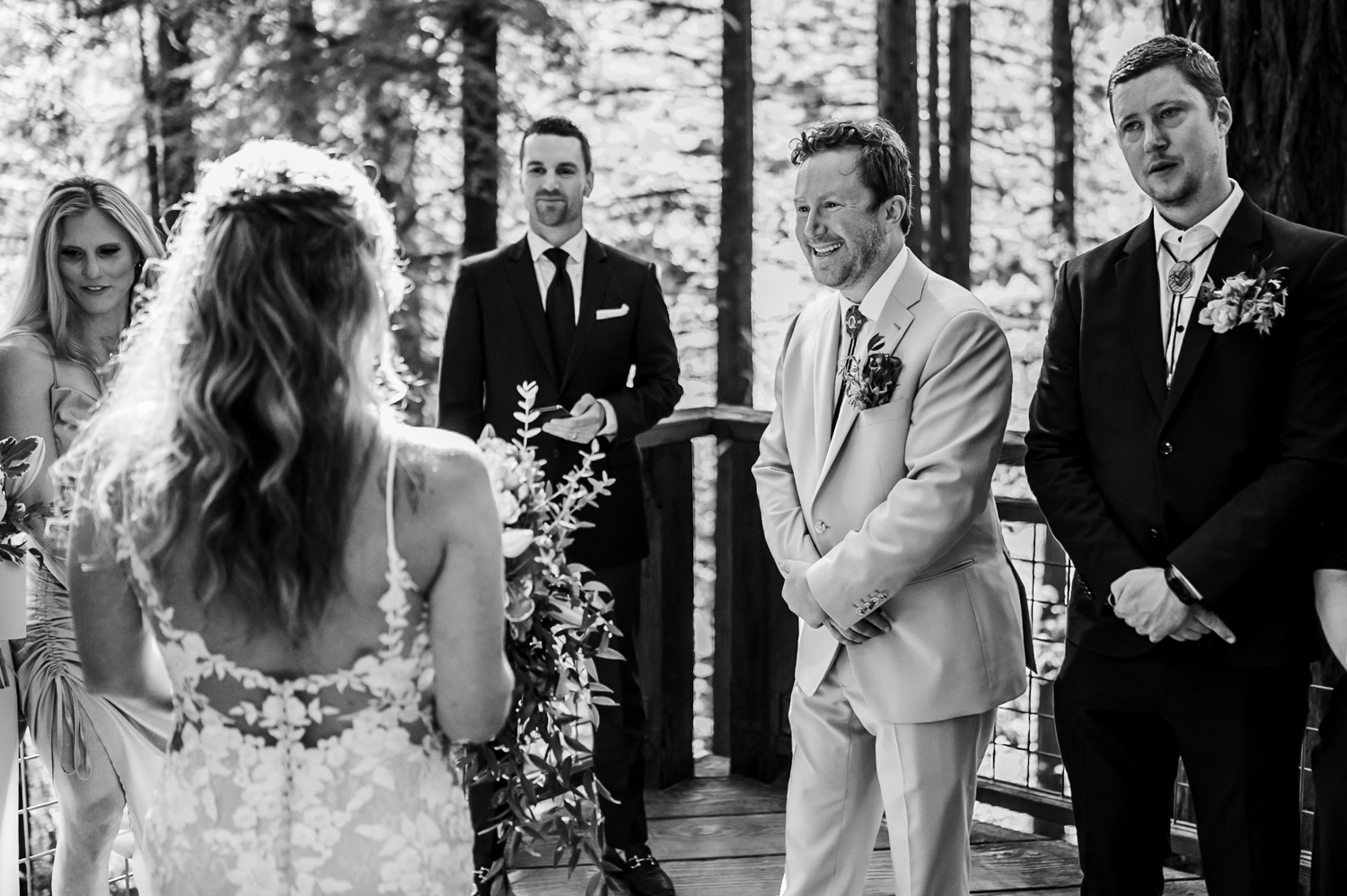 A bride in a lace dress and a groom in a suit stand facing each other, surrounded by guests during their wedding ceremony at the Redwood Deck in Hoyt Arboretums forest setting.