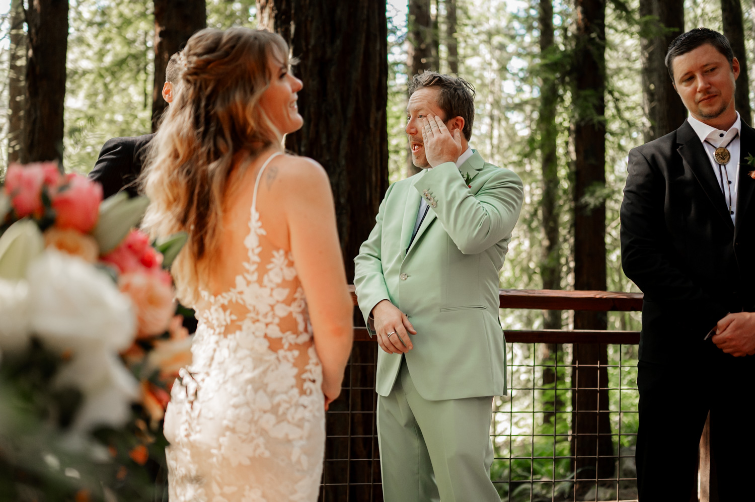 A groom in a light green suit wipes tears from his eyes as he faces his bride during their forest wedding ceremony at the Redwood Deck in Hoyt Arboretum, surrounded by towering trees.