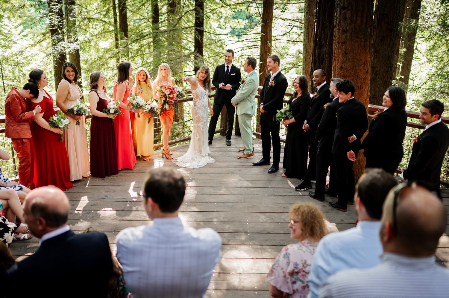 A wedding ceremony at the Redwood Deck in Hoyt Arboretum, featuring the bride and groom encircled by a lively wedding party in colorful clothing.