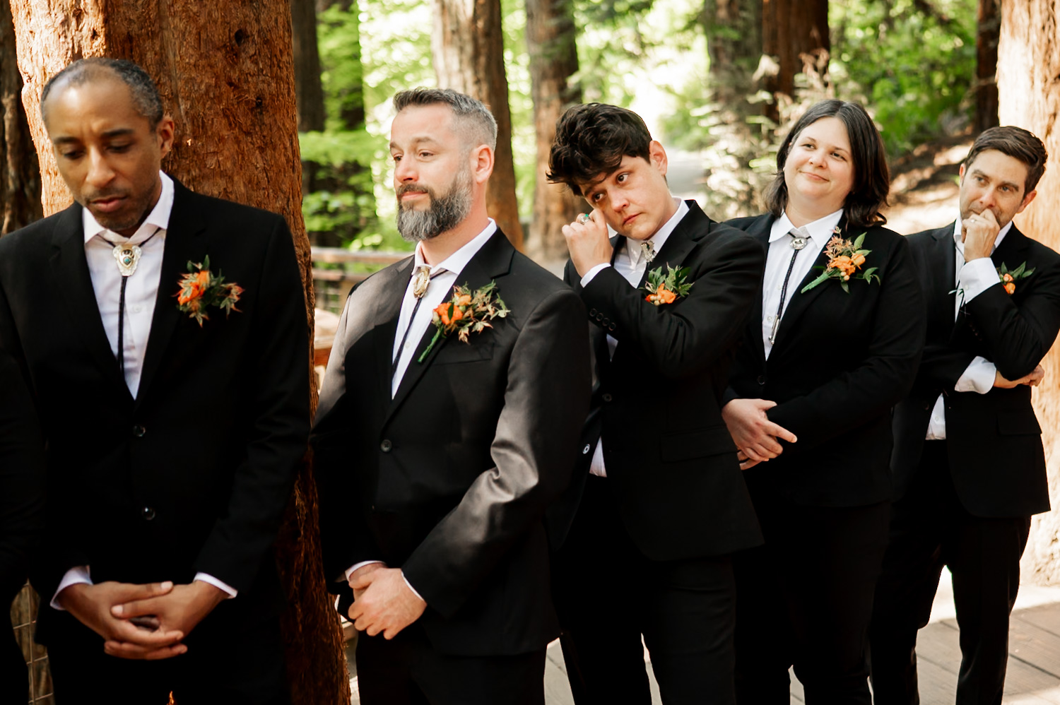 A group of people in suits with orange boutonnieres stands among the tall trees at Hoyt Arboretums Redwood Deck. Some individuals are smiling while one person adjusts their tie, capturing a joyful moment from a wedding ceremony.