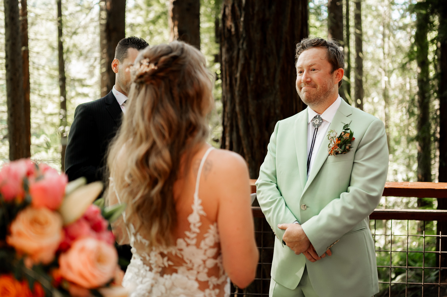 A couple stands amid towering trees at the Redwood Deck in Hoyt Arboretum. The groom wears a light green suit, and the bride holds a bouquet. They exchange vows while being photographed against the backdrop of giant redwoods.