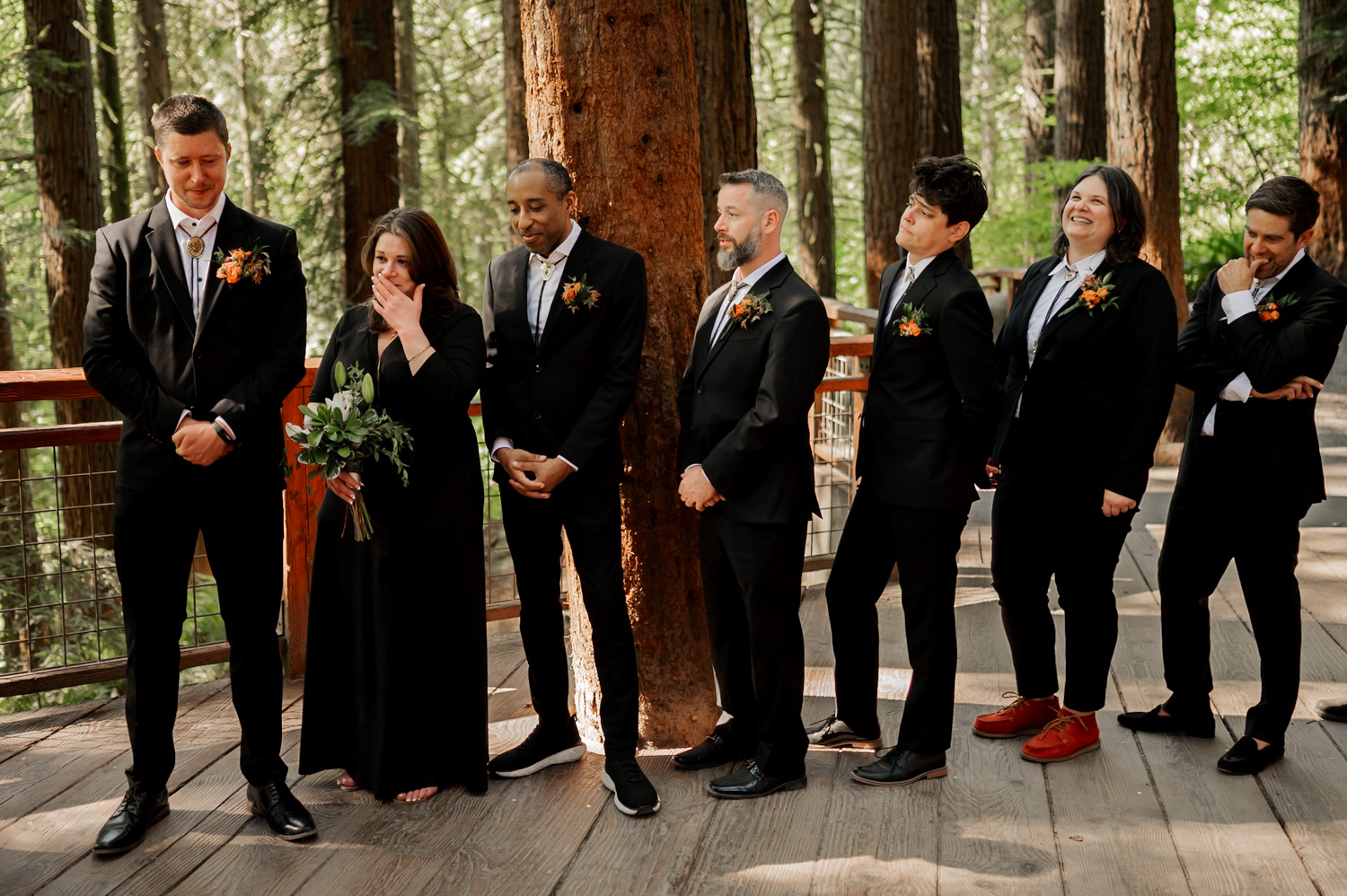 A wedding party in suits and dresses poses joyfully among towering trees at the Redwood Deck in Hoyt Arboretum.