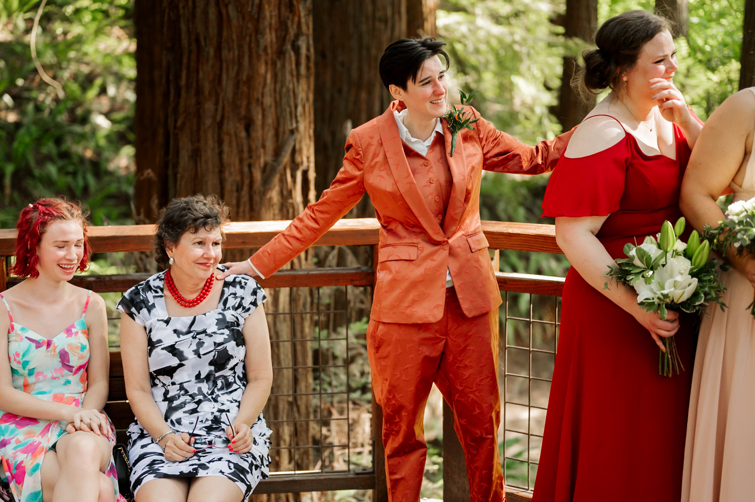 People in vibrant clothing smile and chat amidst a forest backdrop at the Redwood Deck in Hoyt Arboretum during a wedding ceremony.