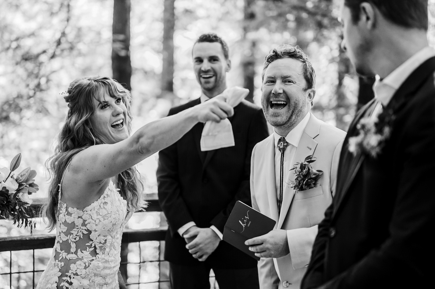 A bride laughing and pointing, standing next to a groom in a white suit, with two men beside them; one man is holding a paper during the wedding ceremony on the Redwood Deck in Hoyt Arboretum.