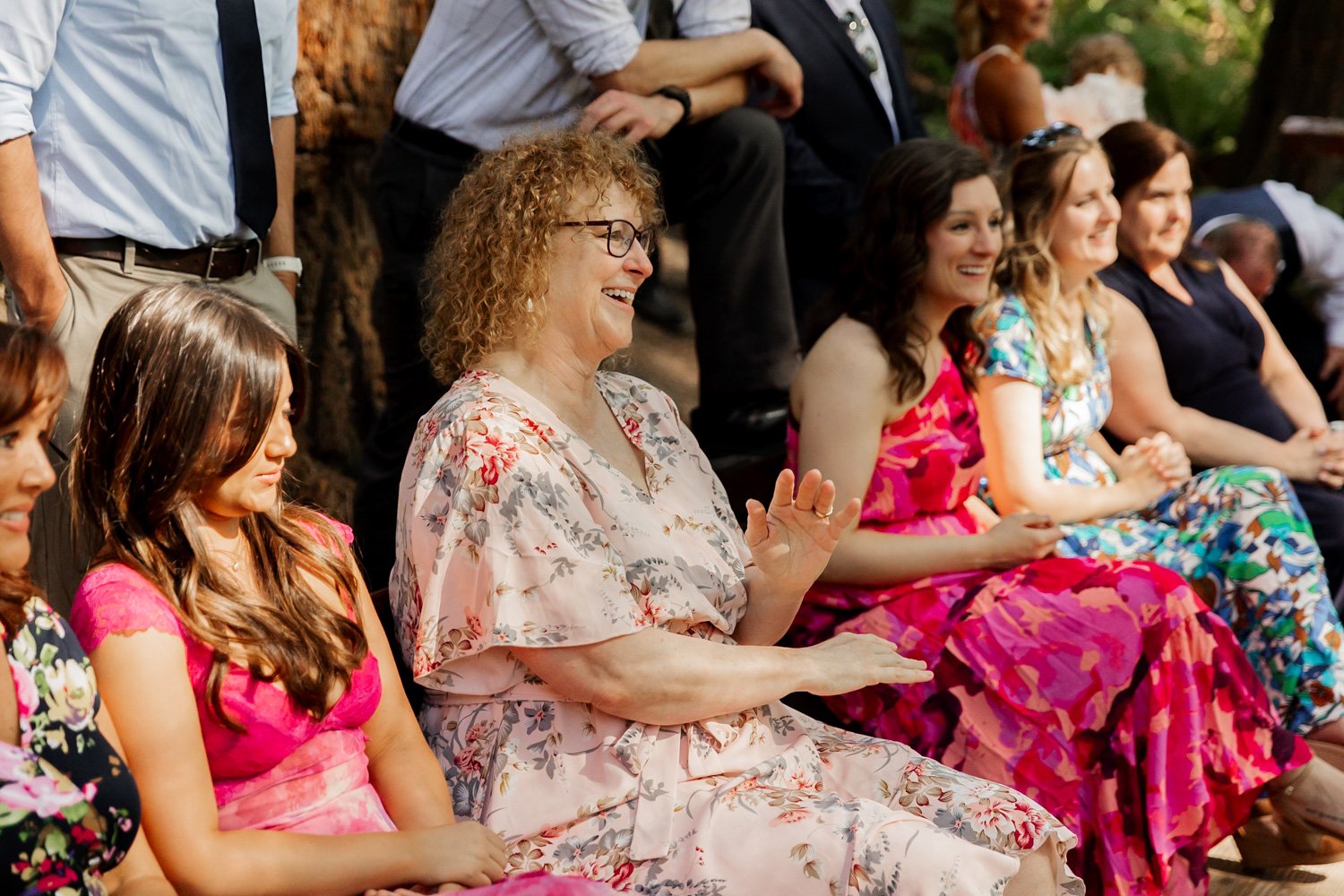 A group of people sitting outdoors at the Redwood Deck in Hoyt Arboretum, smiling and clapping, wearing colorful dresses and shirts during a wedding ceremony.