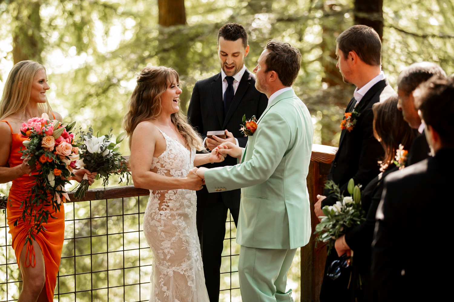 The bride and groom smile during an outdoor wedding ceremony at the Redwood Deck in Hoyt Arboretum, surrounded by attendants and lush greenery.