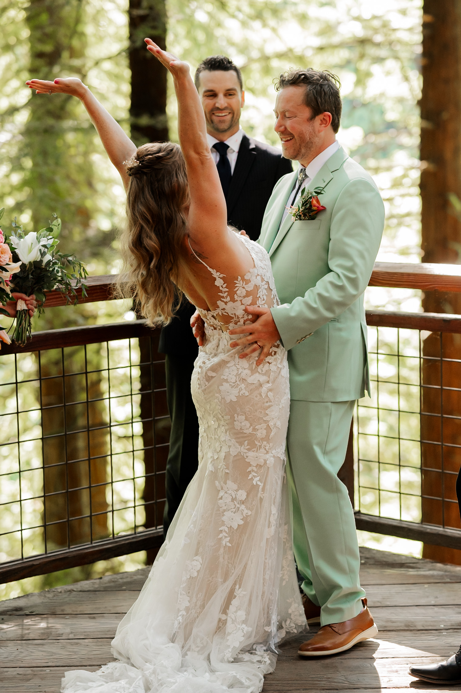 Bride in a lace dress joyfully raises her arms next to the groom in a mint suit and an officiant during their wedding ceremony amidst towering trees at the Redwood Deck, Hoyt Arboretum.