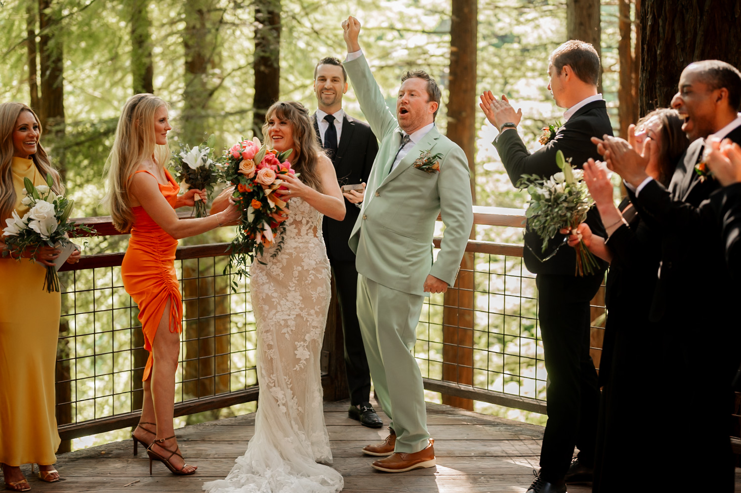 A wedding party celebrates on the Redwood Deck at Hoyt Arboretum, surrounded by tall trees. The groom is cheering energetically while others join in the joyful atmosphere during the ceremony.