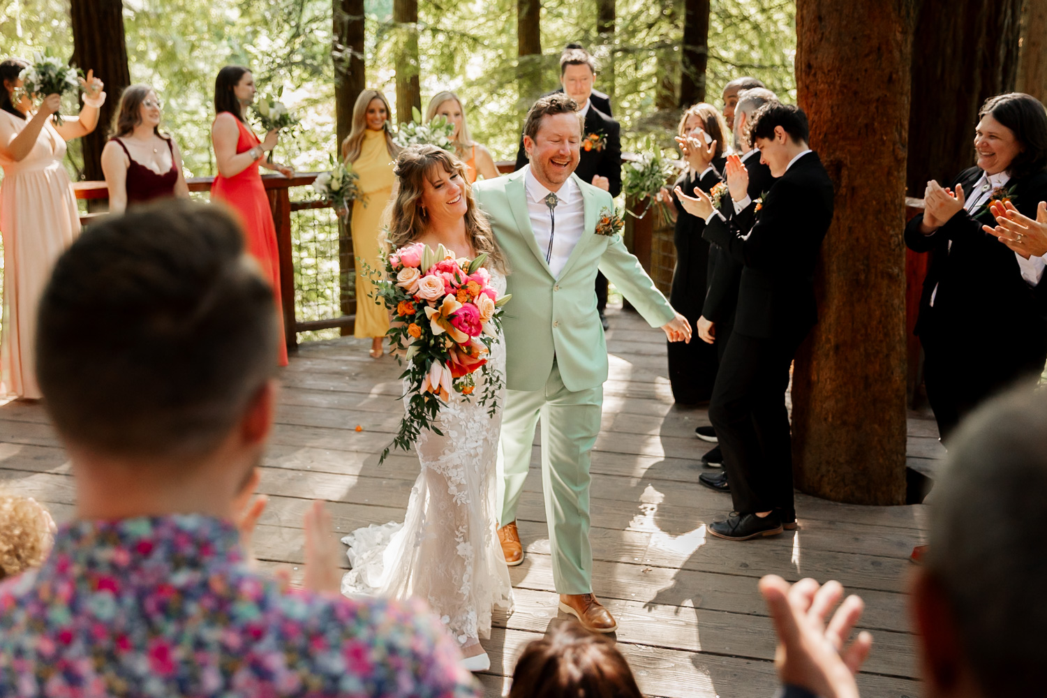 A joyful couple walks hand in hand down the aisle at the Redwood Deck in Hoyt Arboretum, with clapping guests on either side and tall trees surrounding them.