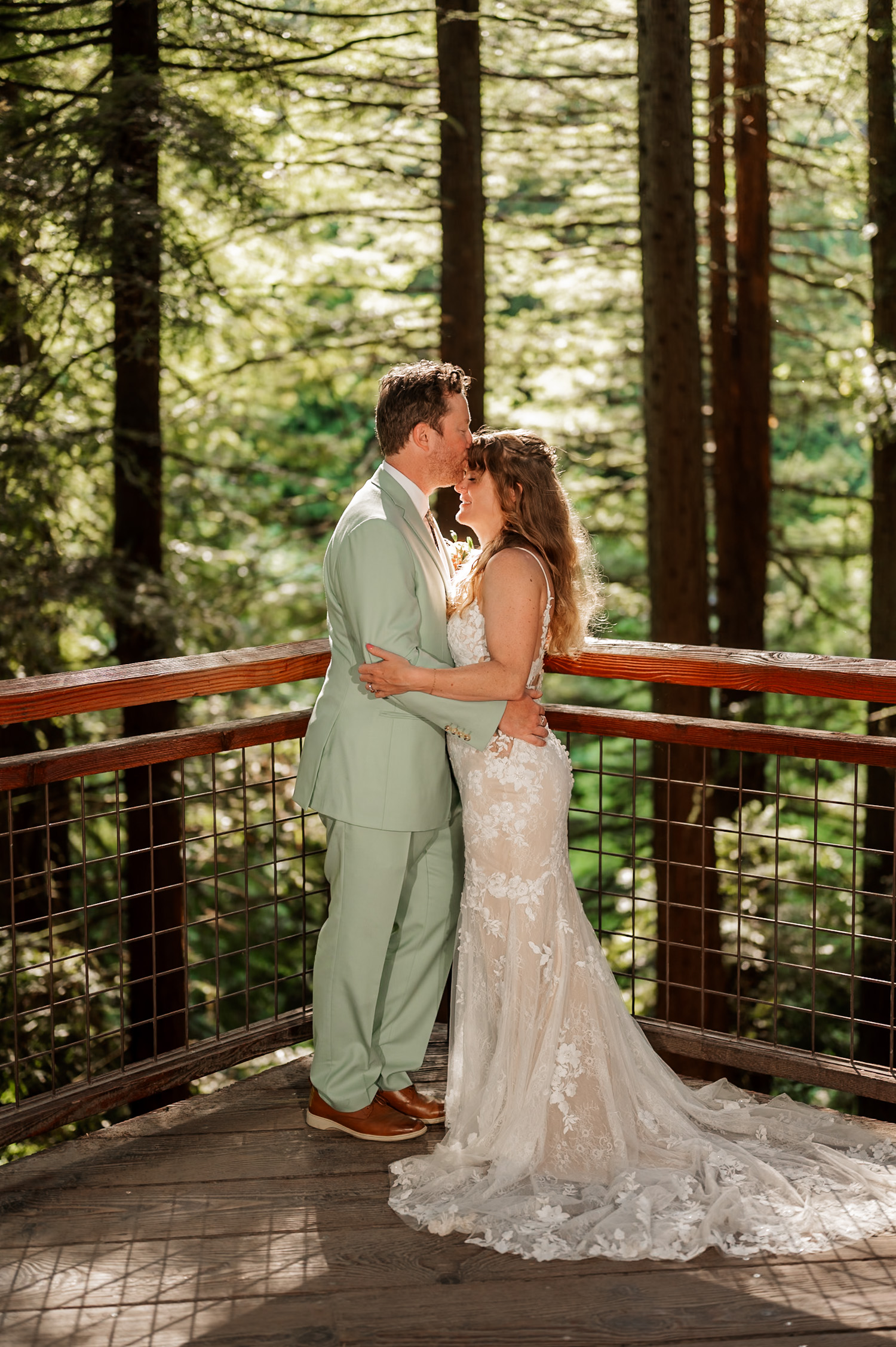 A bride and groom share a kiss on the Hoyt Arboretum Redwood deck with sunlight streaming through the surrounding trees.