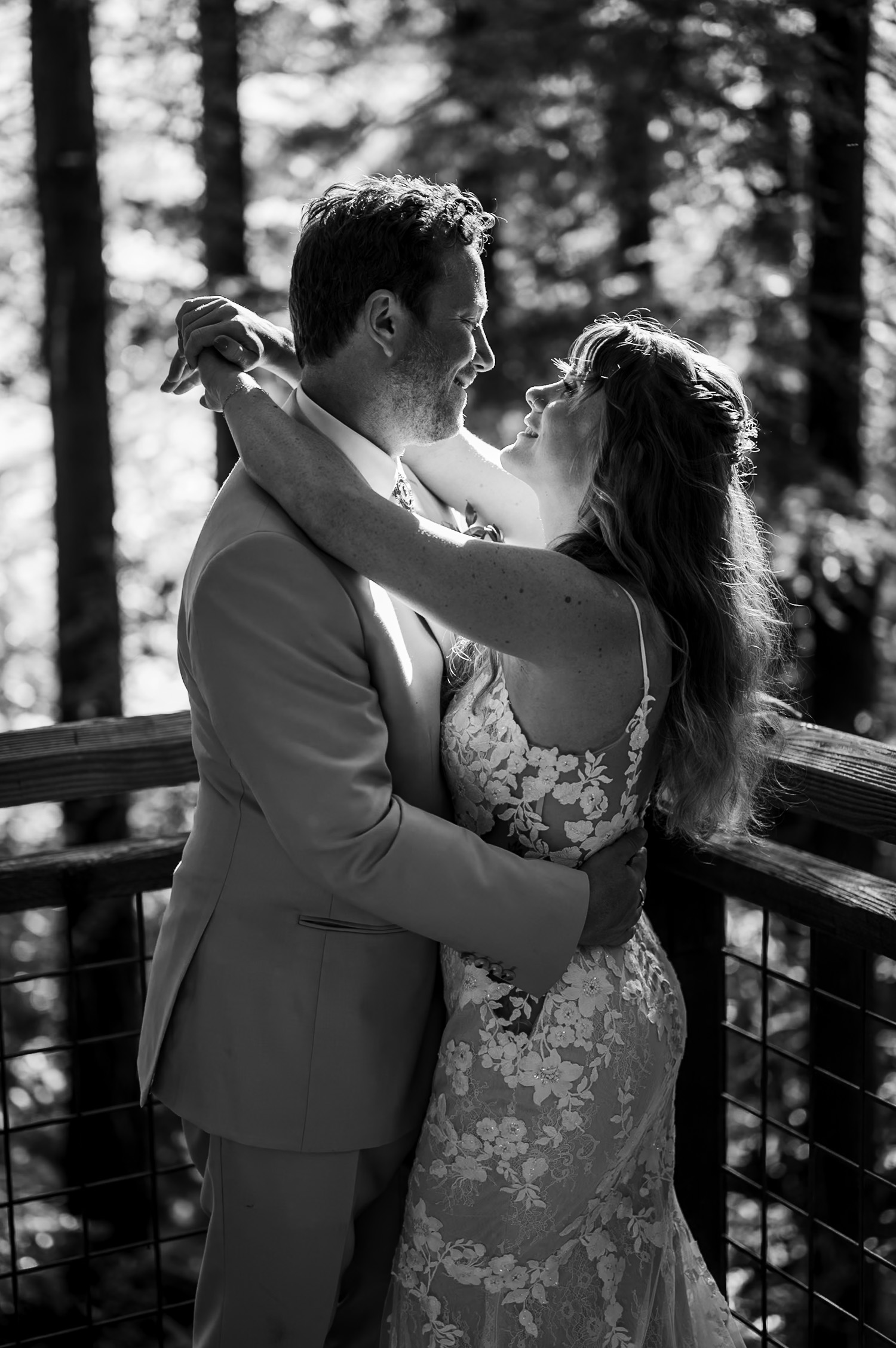 A couple embraces intimately in wedding attire on a sunlit wooden balcony at the Redwood Deck in Hoyt Arboretum, surrounded by lush greenery.