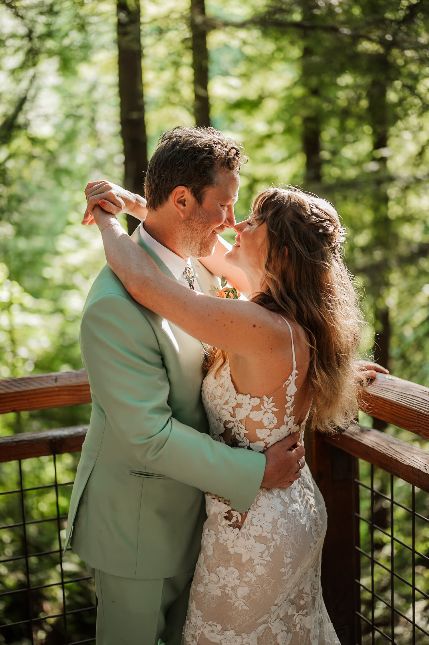 A couple embraces and smiles among towering trees at Hoyt Arboretum, with sunlight filtering through the Redwood deck, creating a picturesque scene.