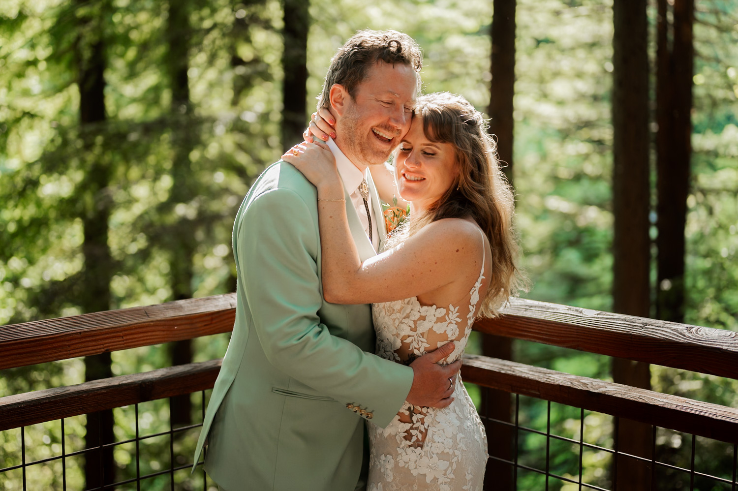 A bride and groom embrace and smile on the Redwood Deck at Hoyt Arboretum, with sunlight streaming through towering trees.