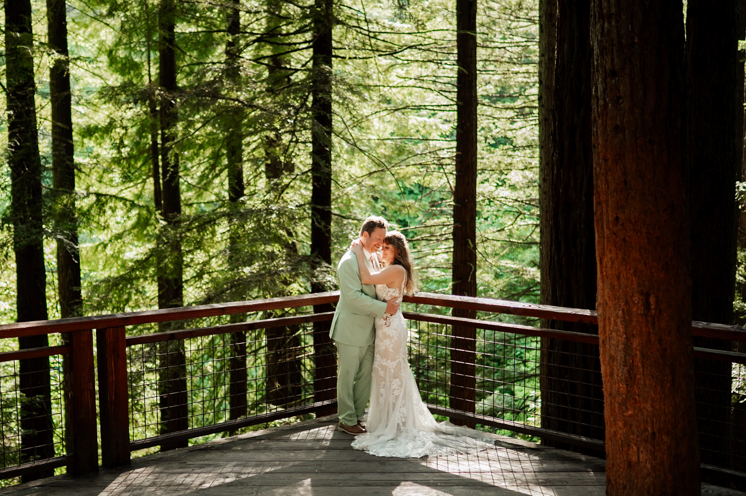 A couple embraces on a wooden platform in Hoyt Arboretum, surrounded by tall trees and dense greenery.