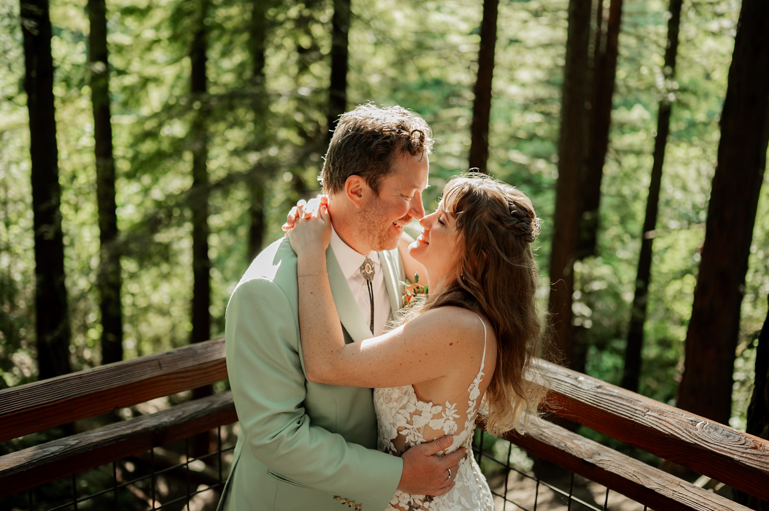 A couple embraces on the Hoyt Arboretum Redwood deck, bathed in sunlight as they share a loving gaze amidst the serene forest setting.