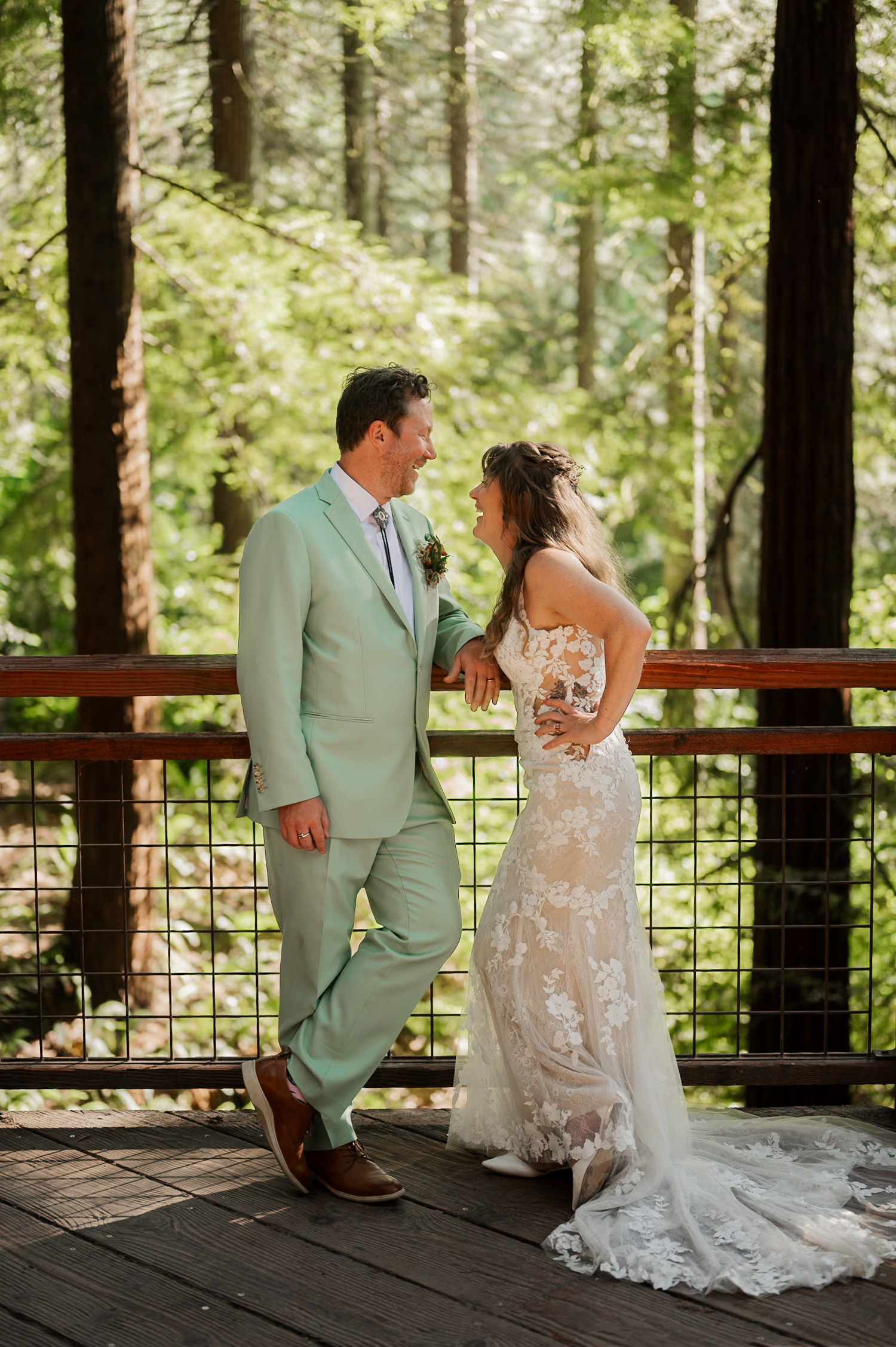 A bride in a lace gown and a groom in a light green suit smile at each other on a forest walkway at Hoyt Arboretum, standing on a redwood deck.