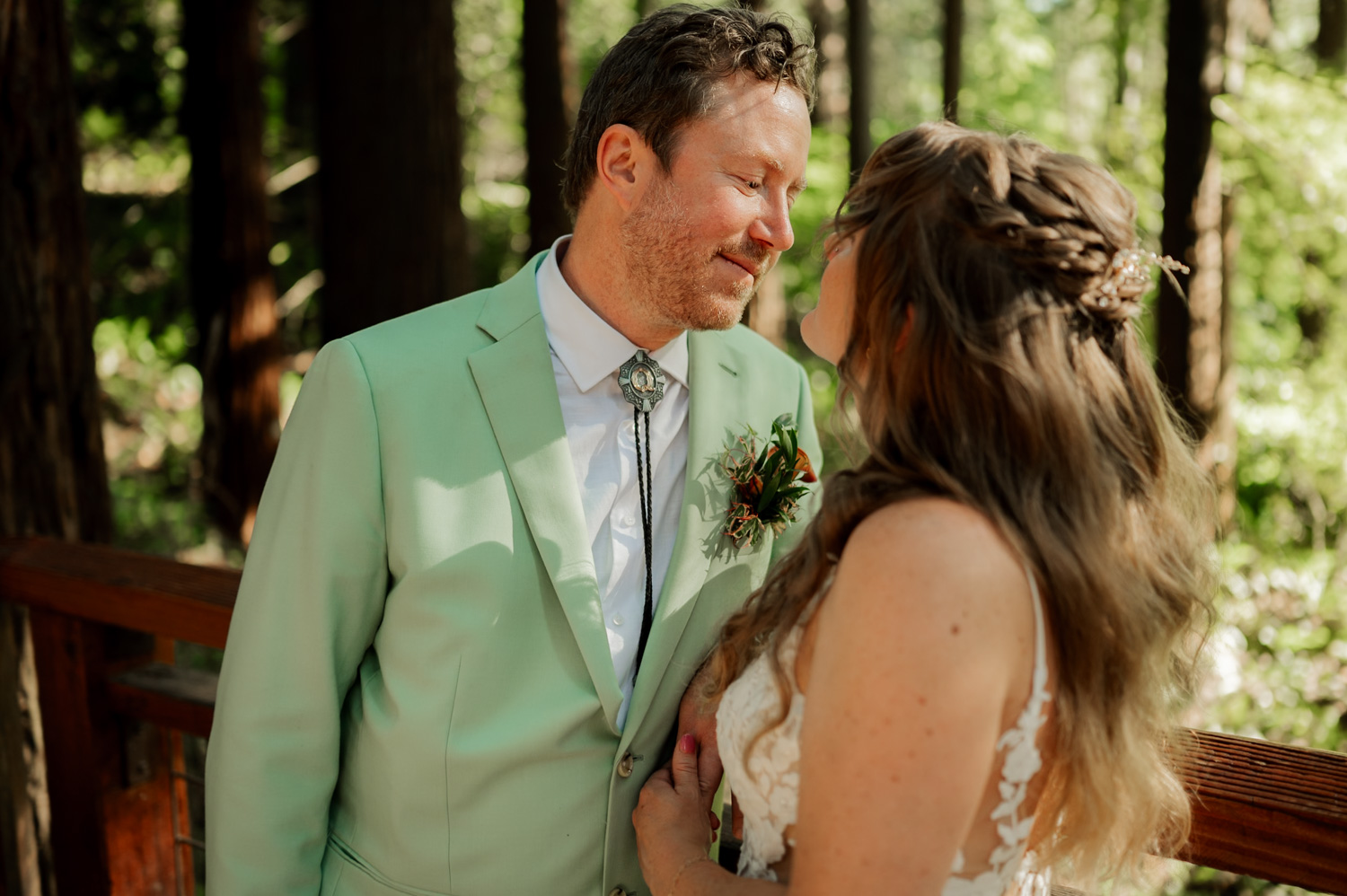 A couple leans toward each other in a forest setting at Hoyt Arboretum, with the man dressed in a light green suit and surrounded by tall trees.