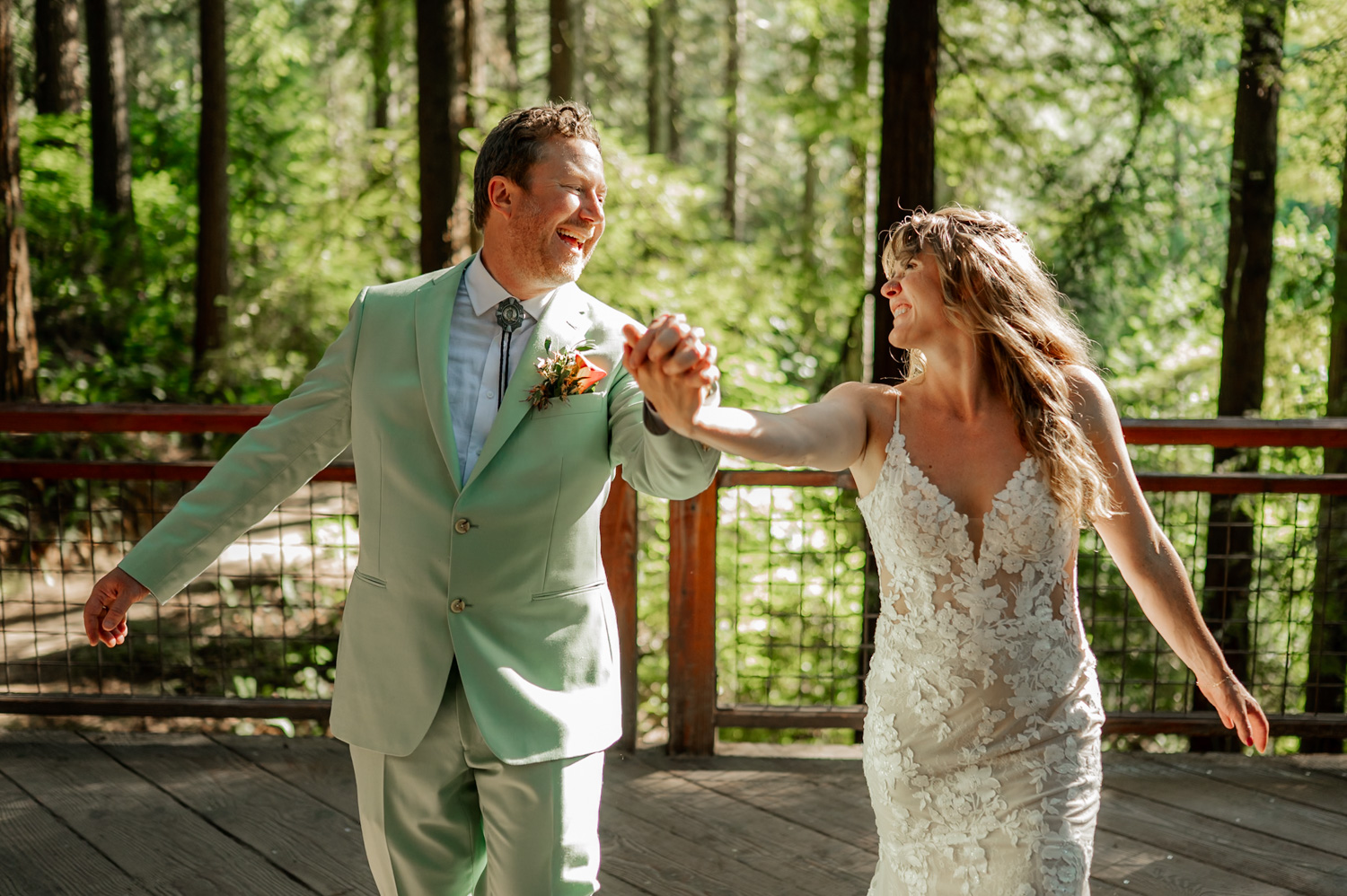 A happy couple in wedding attire holds hands on the Redwood Deck surrounded by the peaceful forest.