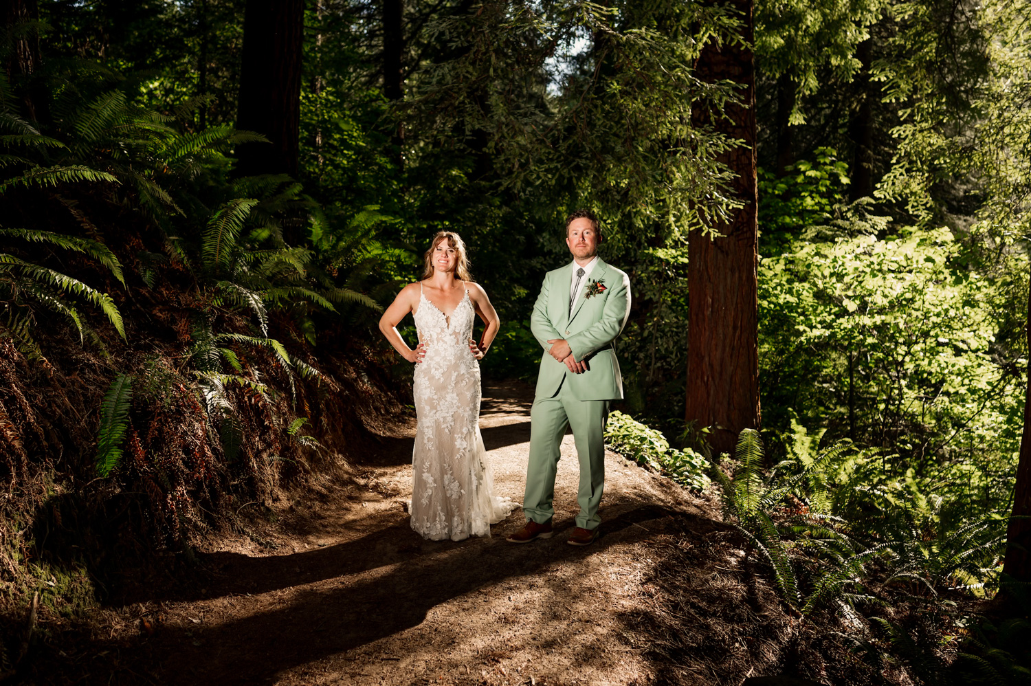 A bride and groom stand confidently in a sunlit forest at Hoyt Arboretum, surrounded by lush green trees and ferns, capturing their love through enchanting Redwood deck wedding photos.