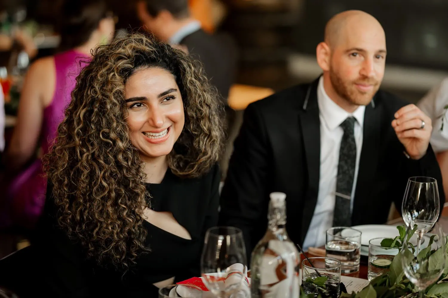 Two people sitting and smiling at a table with glasses and a bottle, enjoying the lively atmosphere of a wedding reception at Coopers Hall.