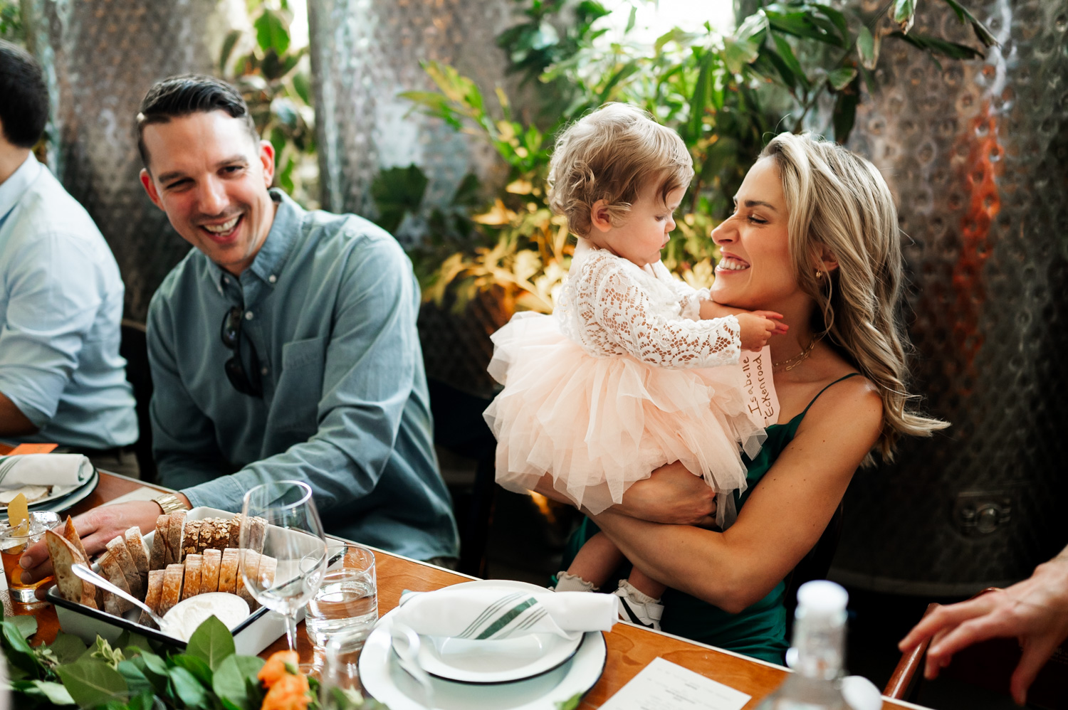 A woman smiling at a child in a dress at a dining table, with a man seated nearby and plants in the background, during a wedding reception at Coopers Hall.