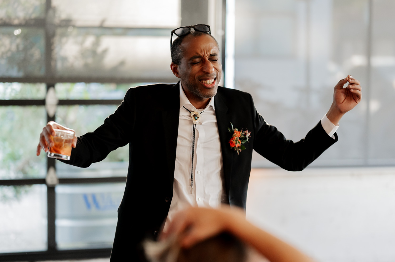 A man in a suit dances with a drink in hand and sunglasses perched on his head, eyes closed, enjoying the moment at a wedding reception in Coopers Hall. The room is brightly lit.