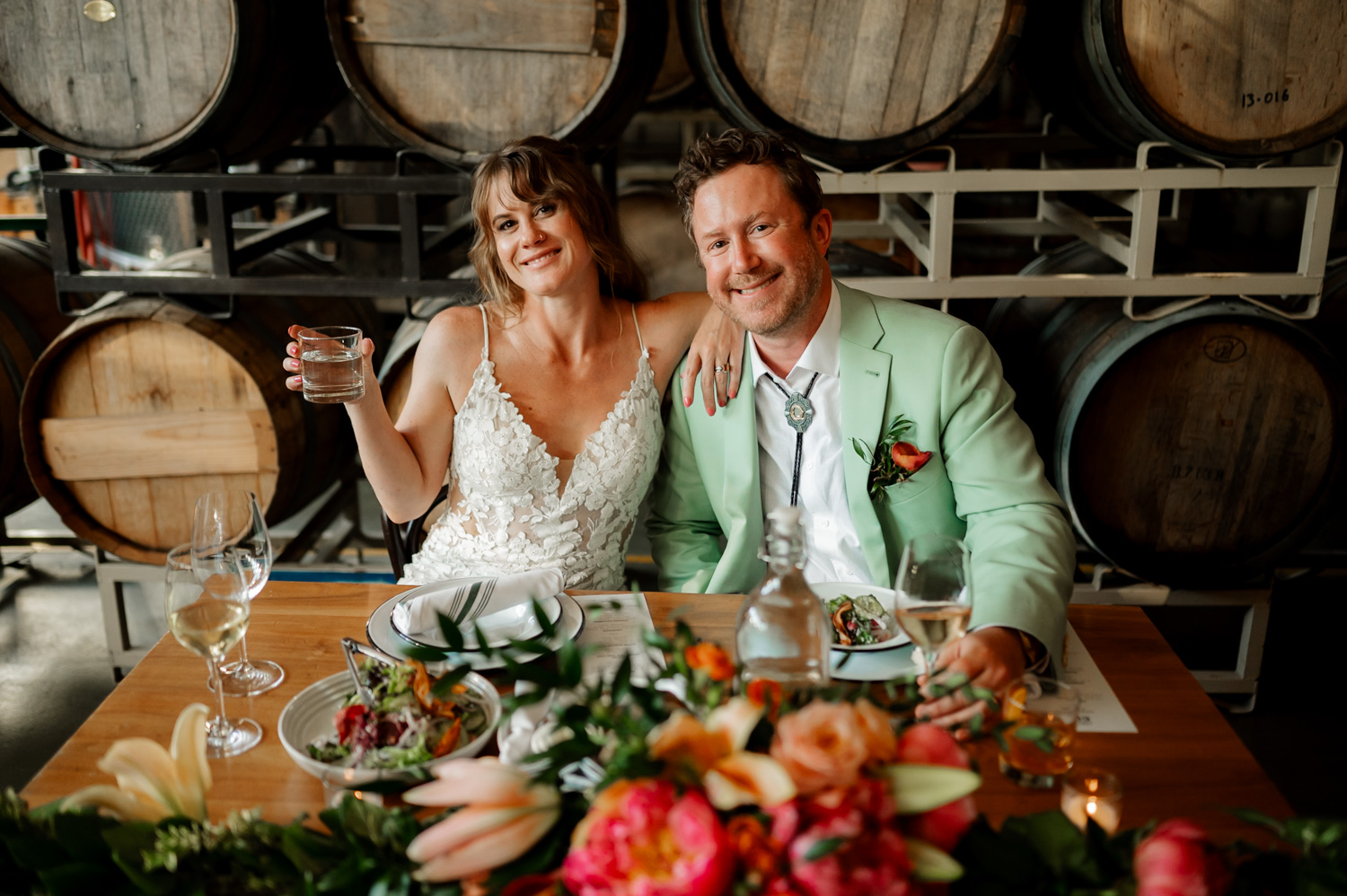 A smiling couple in wedding attire sits at a decorated table with barrels in the background, celebrating their wedding reception at Coopers Hall.