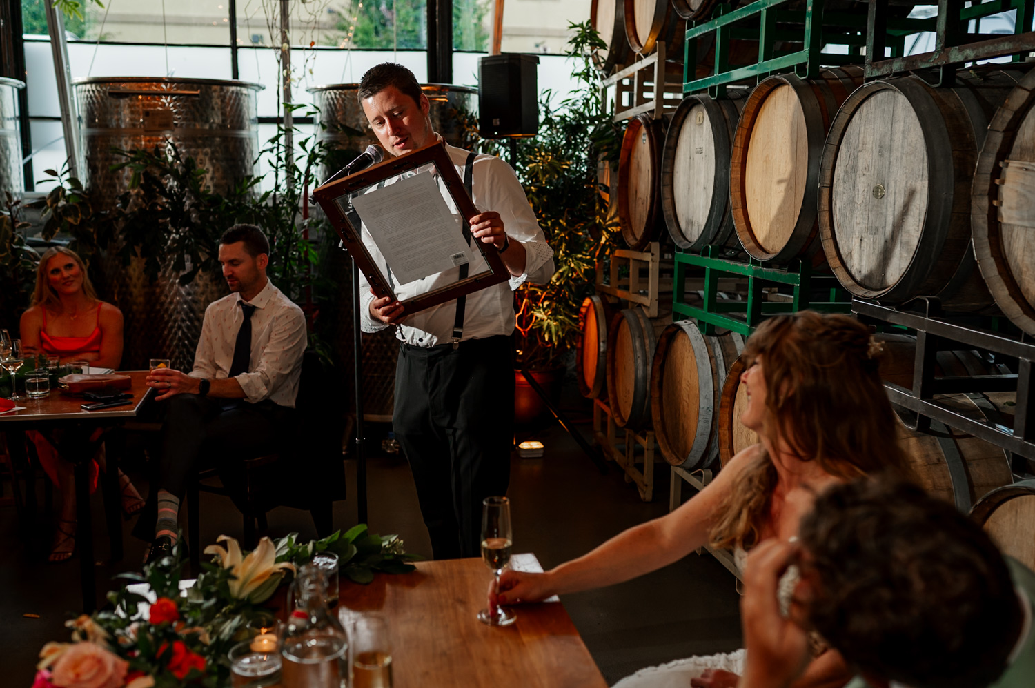 A man gives a speech while holding a framed document at a wedding reception, with wine barrels in the background at Coopers Hall.