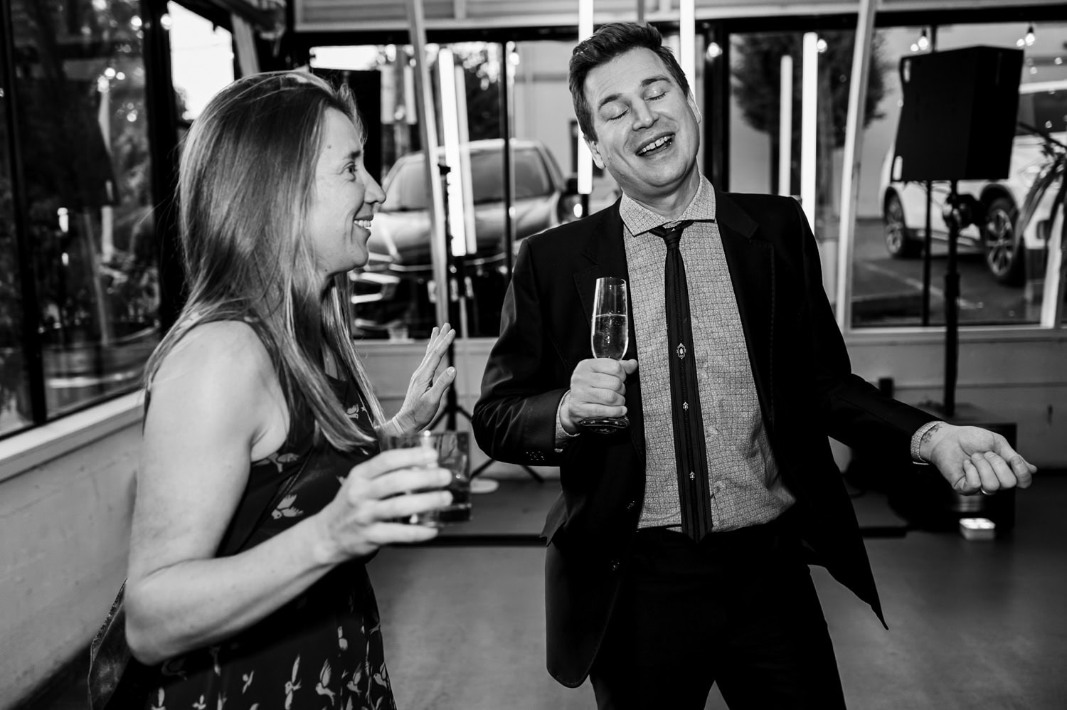 Two people dressed in formal attire, laughing and holding drinks in a brightly lit room with large windows during a wedding reception at Coopers Hall.